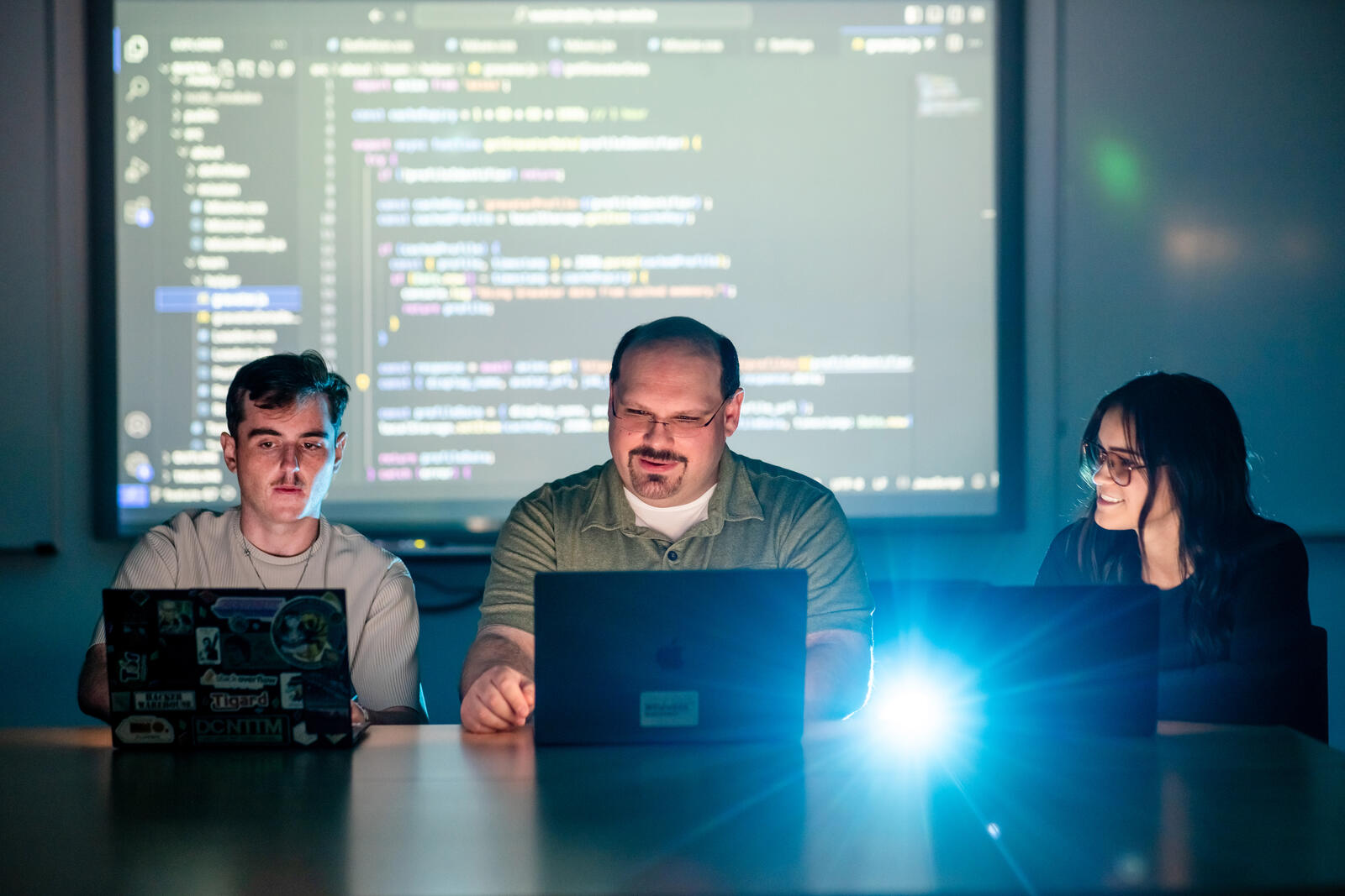 Three C3 Lab members working at laptops in a darkened room, with a large code editor projected on the screen behind them